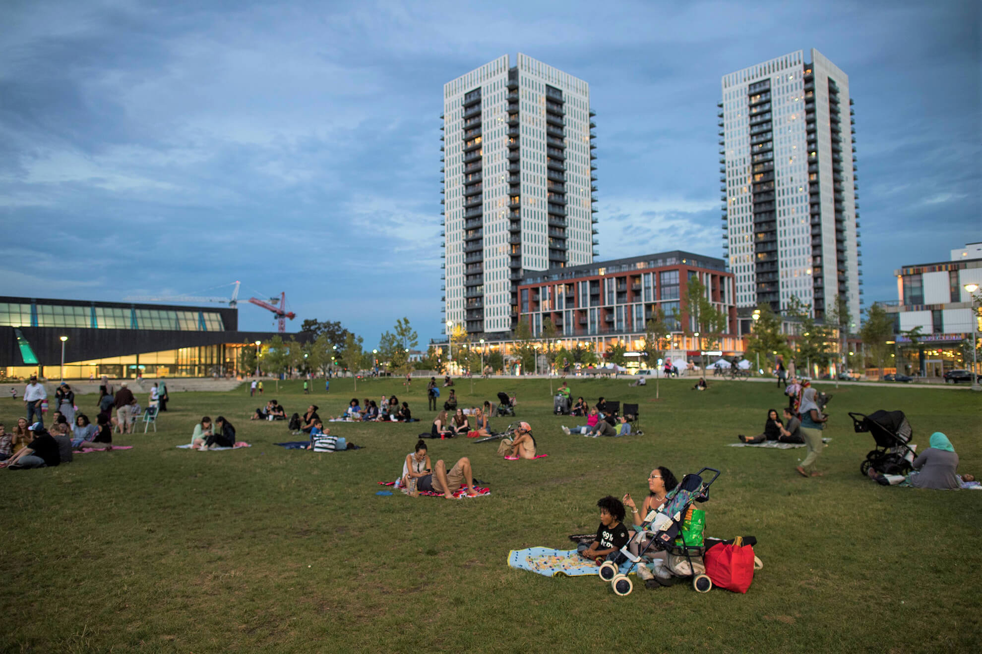 Different groups of people in a park. Most are sitting with family enjoying the day. Towers in the background