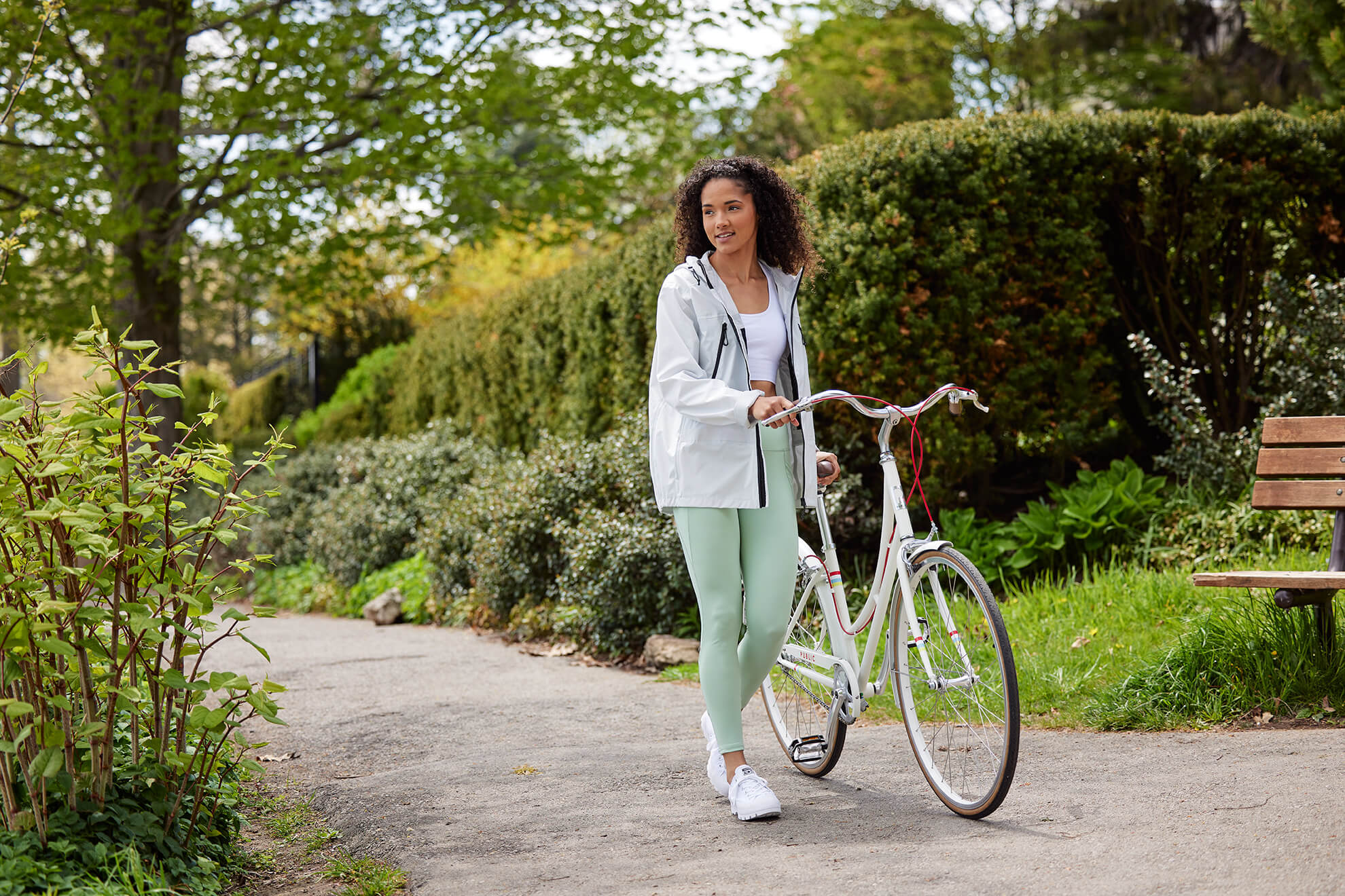 Woman holding her bike on a bike path. Standing in front of a bench.